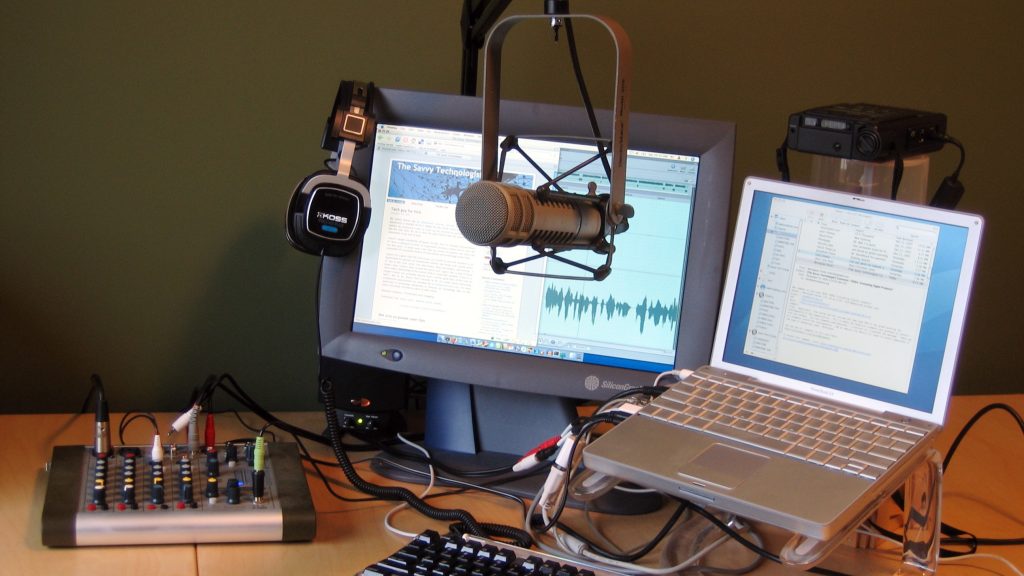 Home voice recording setup with a hanging microphone, headphones, small mixer and an audio waveform on the monitor