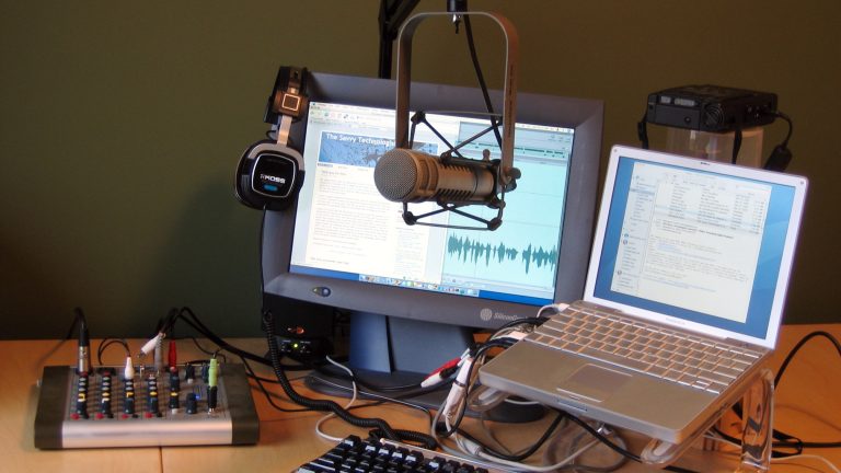 Home voice recording setup with a hanging microphone, headphones, small mixer and an audio waveform on the monitor